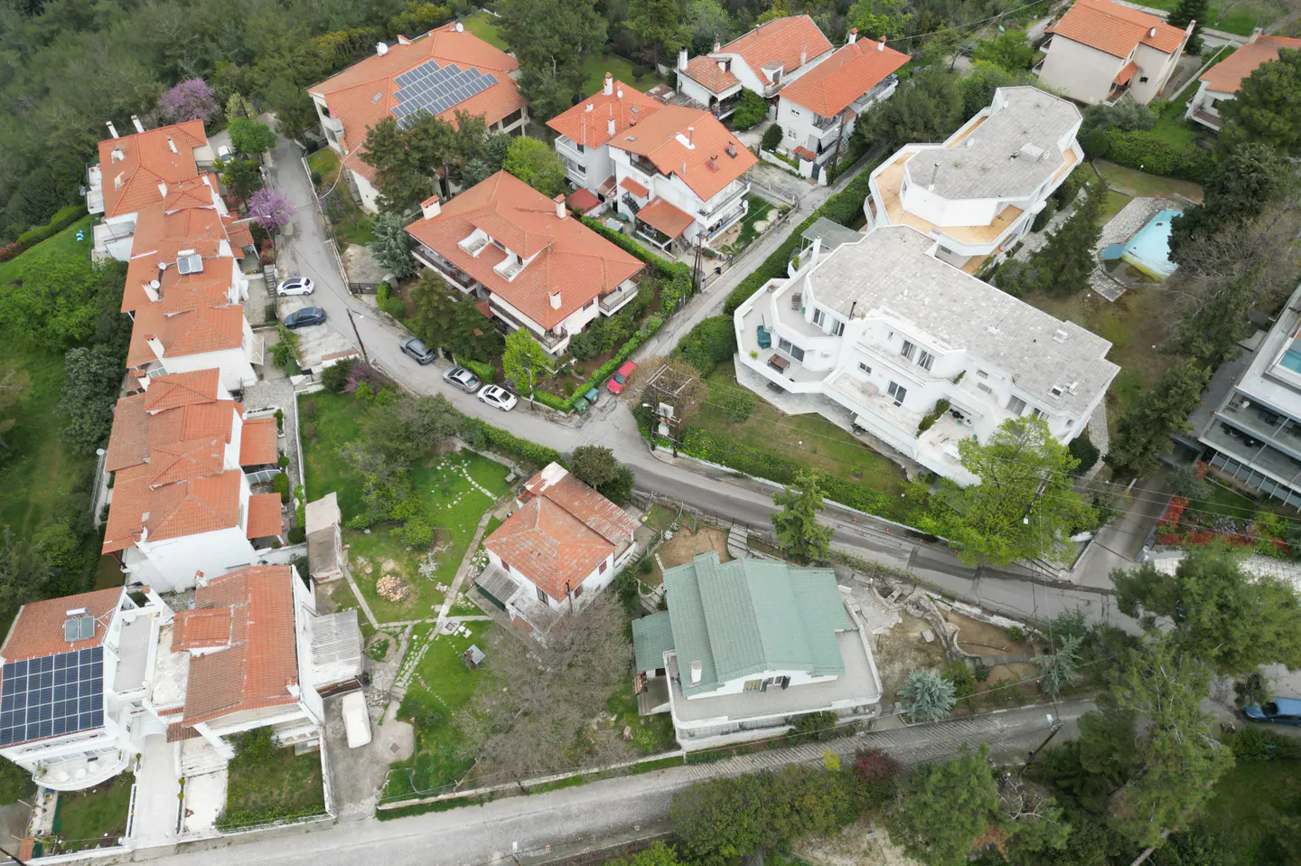 Aerial view of a residential neighborhood with houses, trees, and winding roads. Some houses have red tile roofs and solar panels.