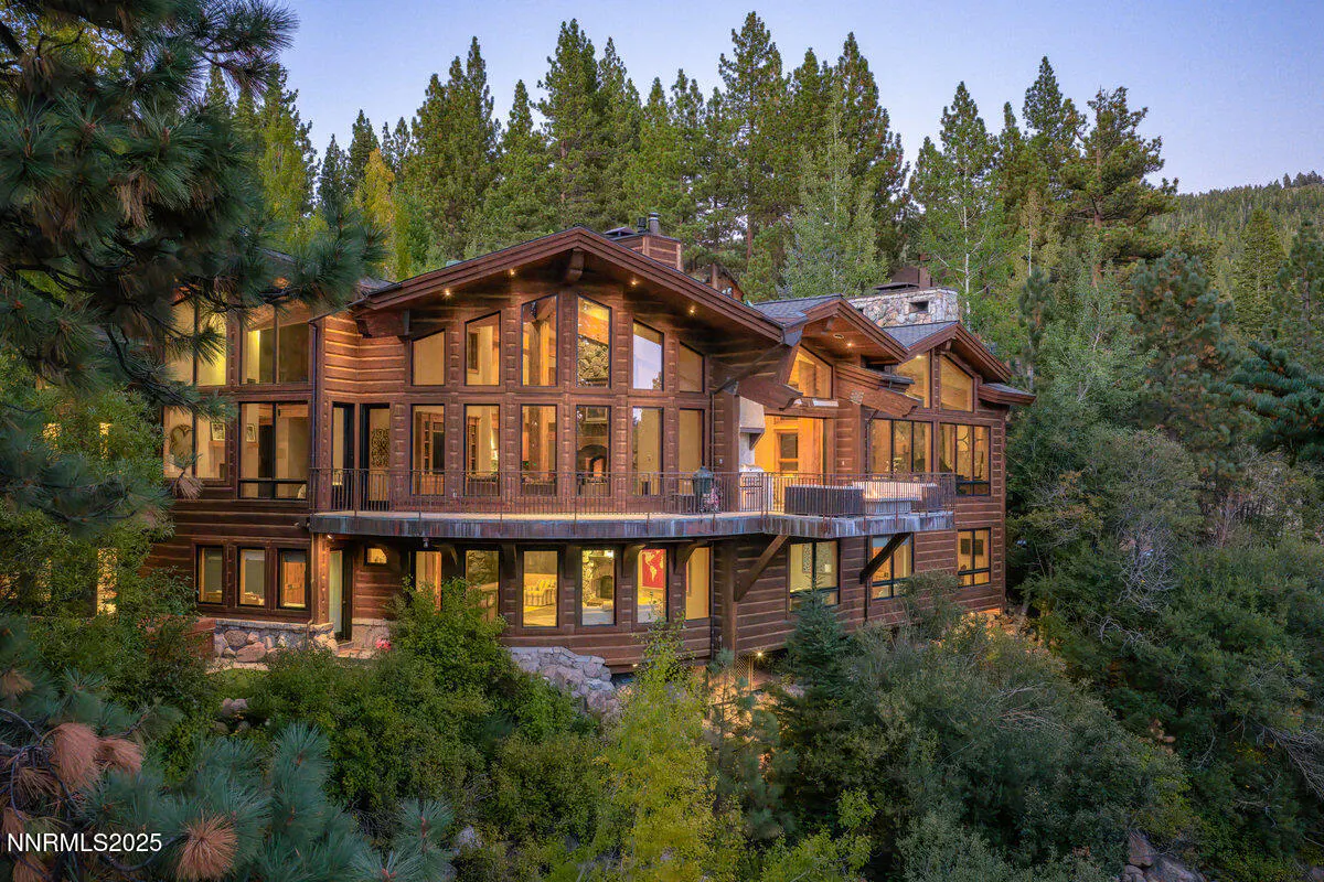 A large, multi-story log cabin with many windows sits nestled among tall green trees.