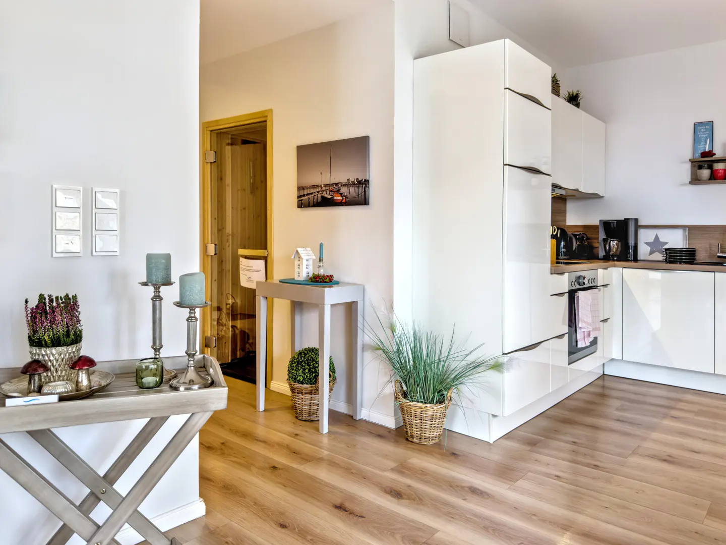 Bright apartment interior with wood floors, white walls, and a modern kitchen. A sauna door is visible in the background.