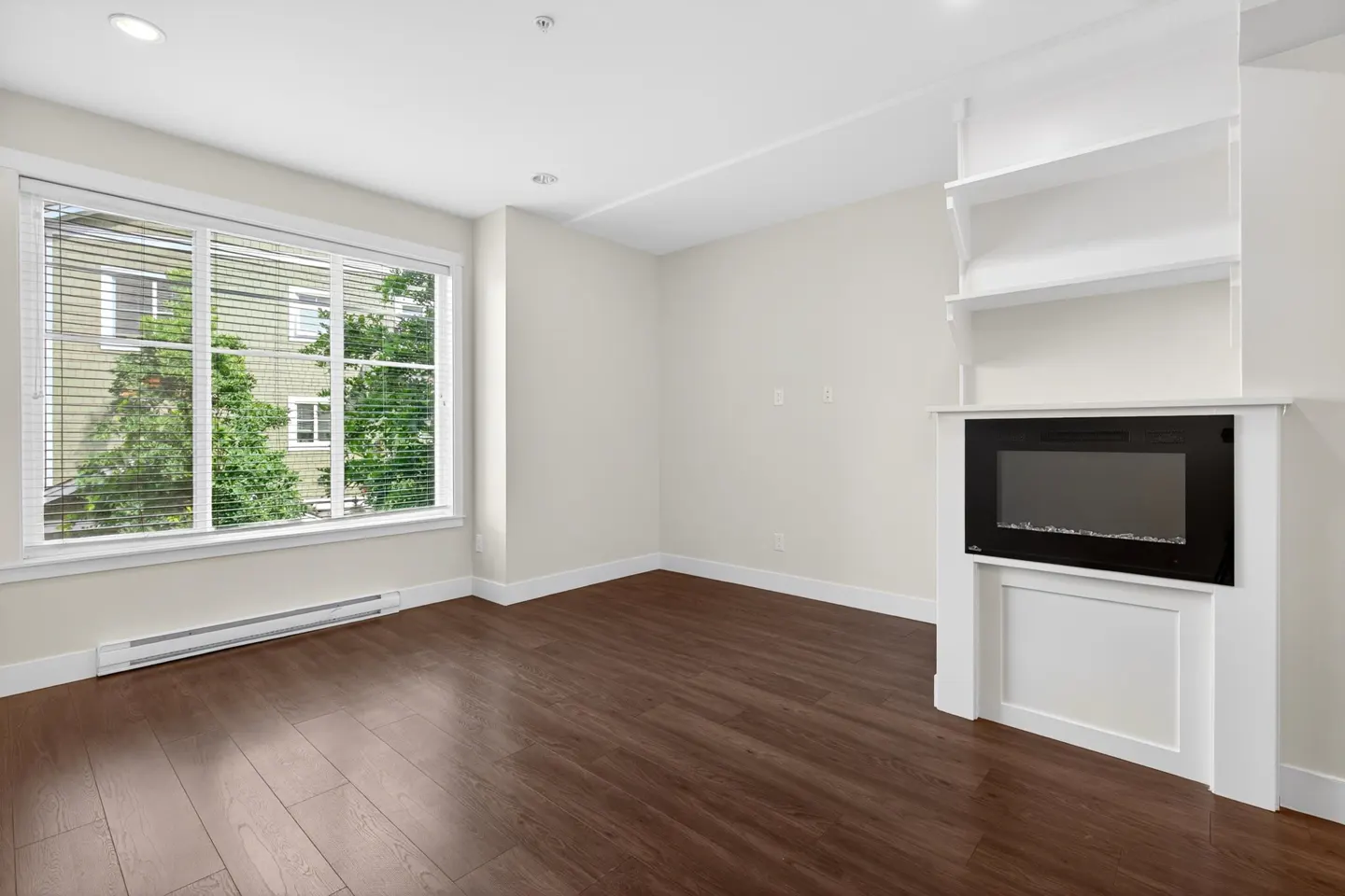 Bright room with dark wood floors, a large window with blinds, and a white fireplace with built-in shelving above.