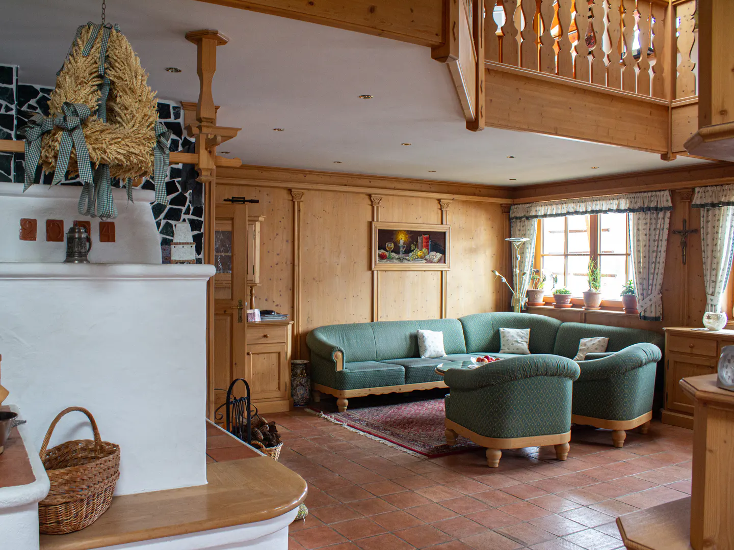 Rustic living room with wood-paneled walls, green sectional sofa, tiled floor, and a white masonry stove with a wheat wreath.