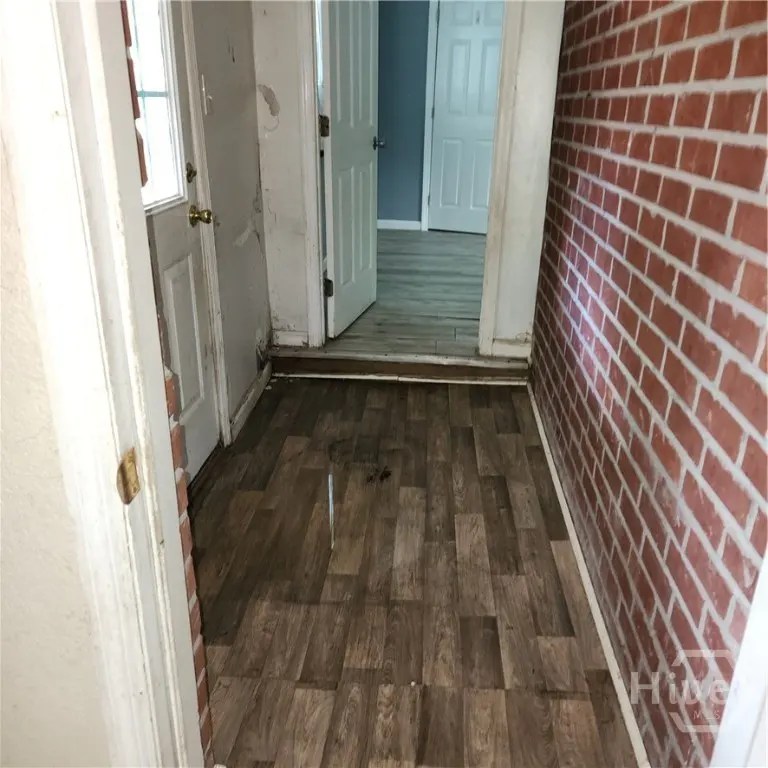 Hallway with wood-look flooring, brick accent wall, and white doors. The walls are white and show signs of wear.