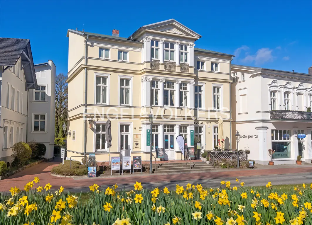 Three-story yellow building with white trim and many windows, seen from across the street with yellow daffodils in the foreground.