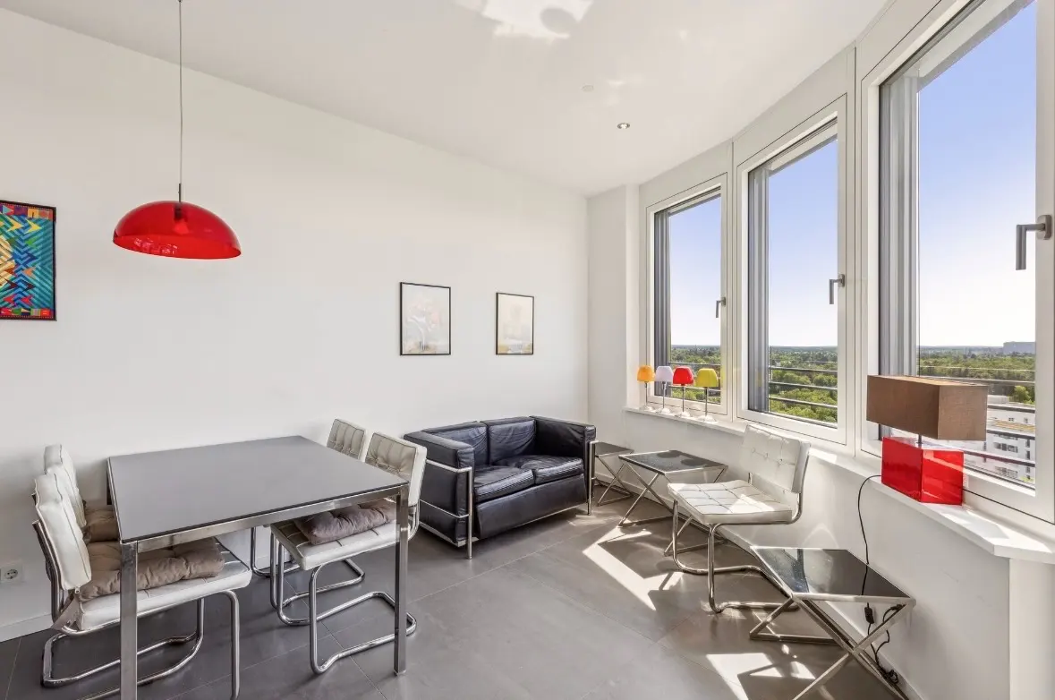 Modern living room with a dining table, black leather sofa, white chairs, red pendant light, and large windows with a view.