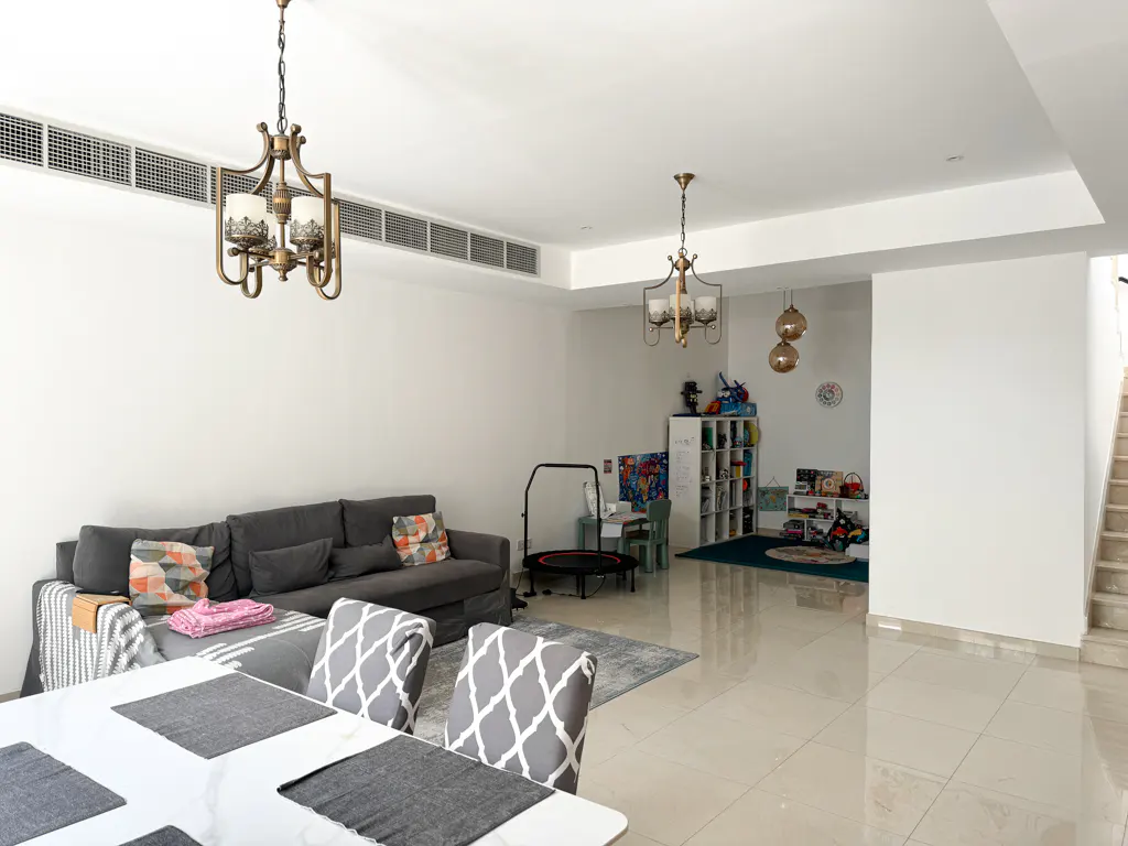 Bright living room with gray sofa, patterned chairs, and a child's play area with toys and a trampoline. Two chandeliers hang from the white ceiling.
