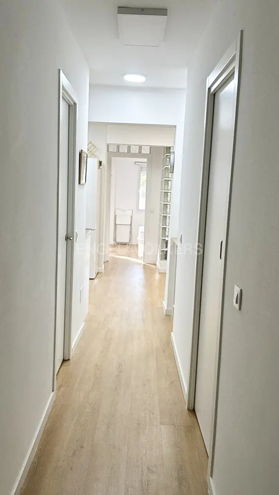 Bright hallway with light wood flooring, white walls, and closed doors leading to a sunlit room in the background.