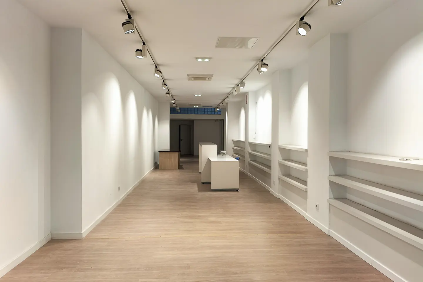 Bright, empty retail space with white walls, wood floors, and track lighting. Shelving units line one wall.