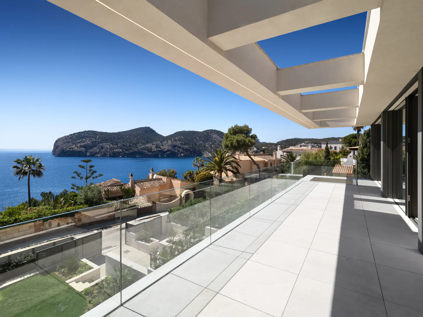 Balcony view of ocean and island. White tiled floor, glass railing, and modern white pergola. Blue sky.