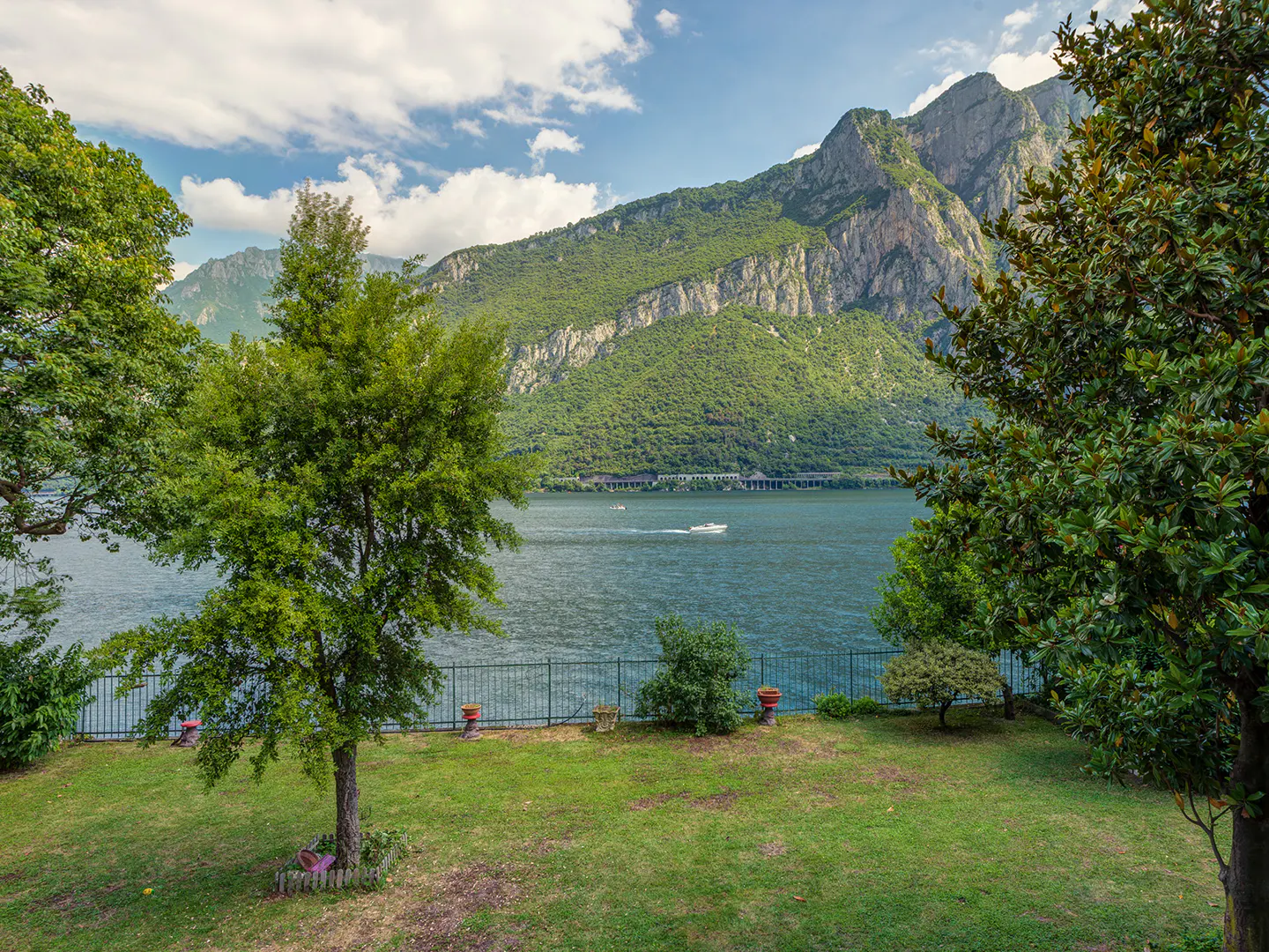Lush green lawn with trees overlooking a blue lake and mountains under a partly cloudy sky. A boat is on the water.