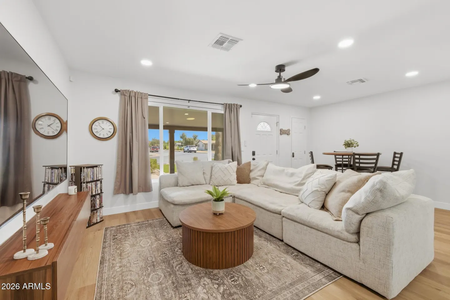 Bright living room with a large white sectional sofa, round wood coffee table, and patterned rug. A dining table and chairs are visible in the background.