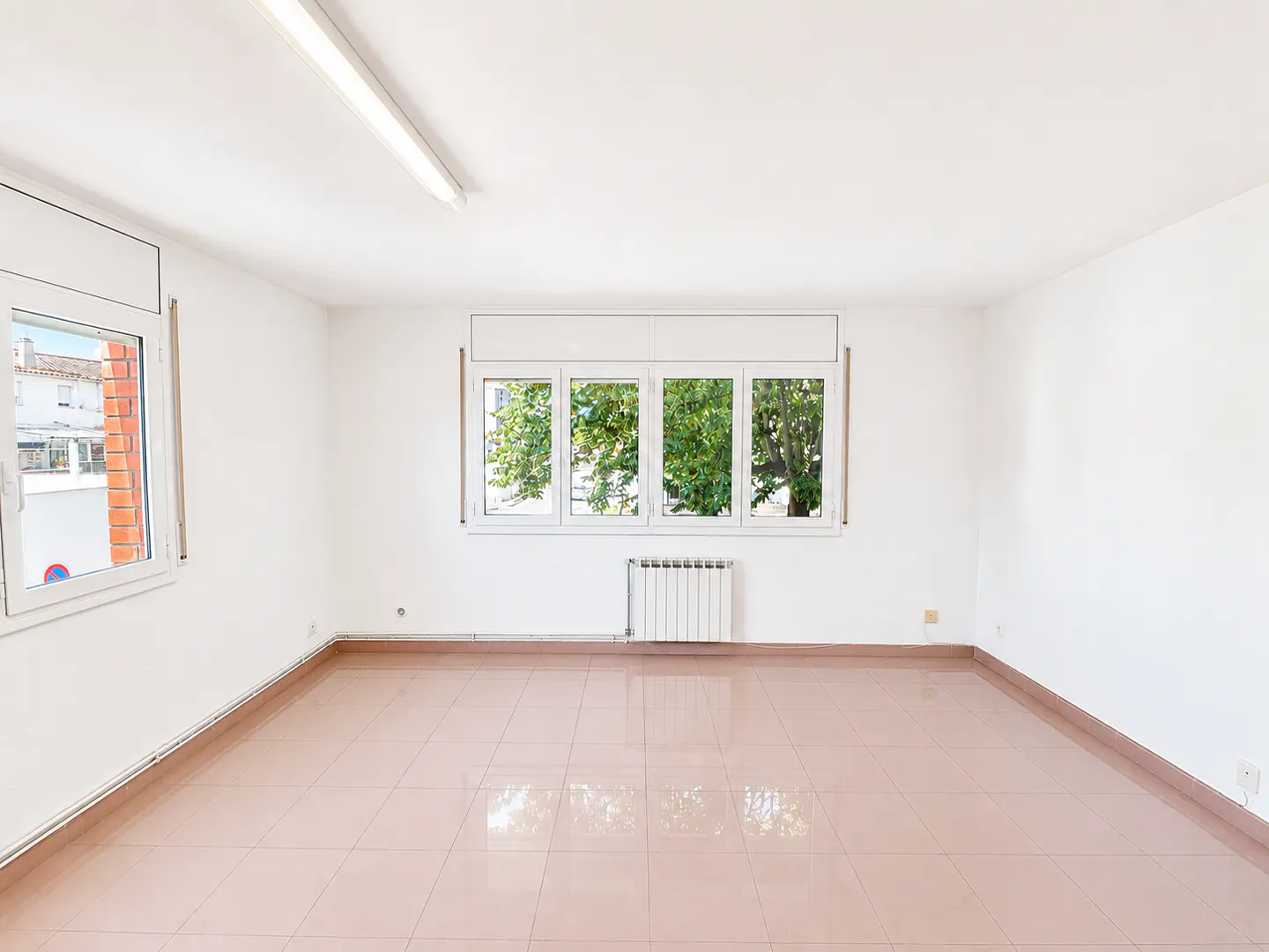 Empty room with white walls, light-colored tiled floors, a radiator, and windows looking out onto green trees.