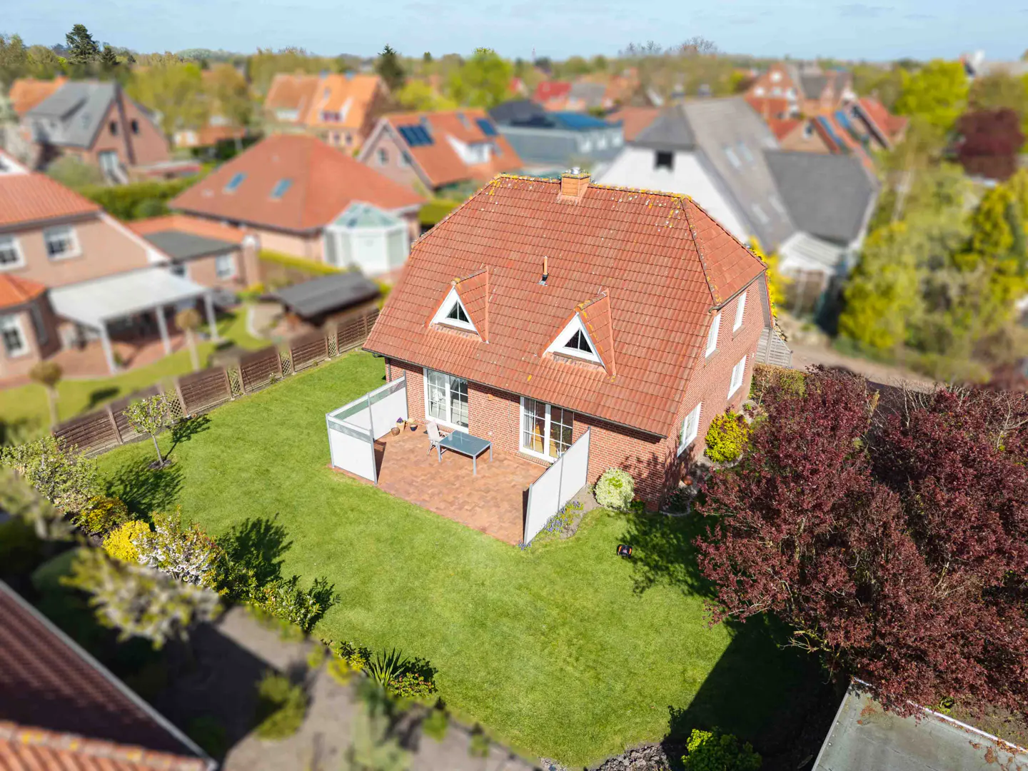 Aerial view of a red brick house with a red tile roof, a patio, and a green lawn in a suburban neighborhood.
