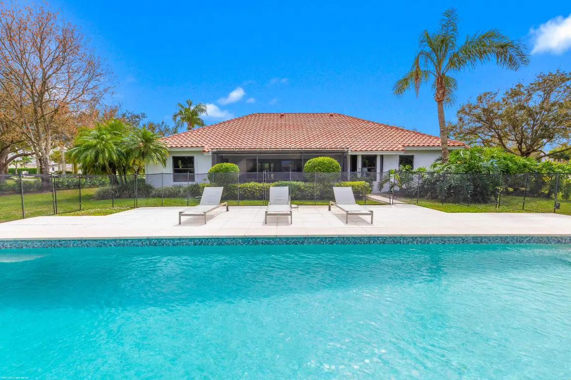 A backyard pool with three lounge chairs on a white patio, a white house with a red tile roof, and a blue sky.