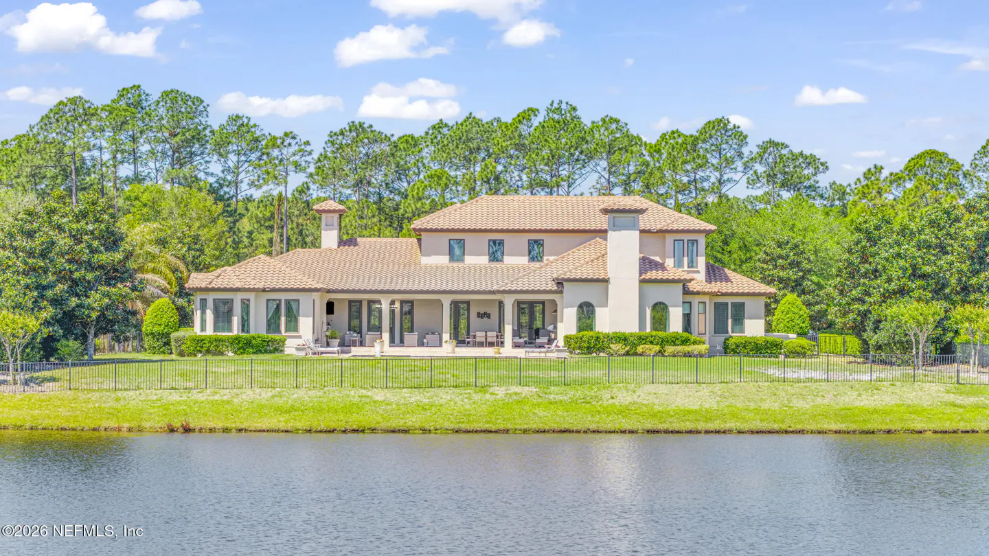 A large, beige house with a brown tile roof sits on a green lawn next to a lake under a blue sky.