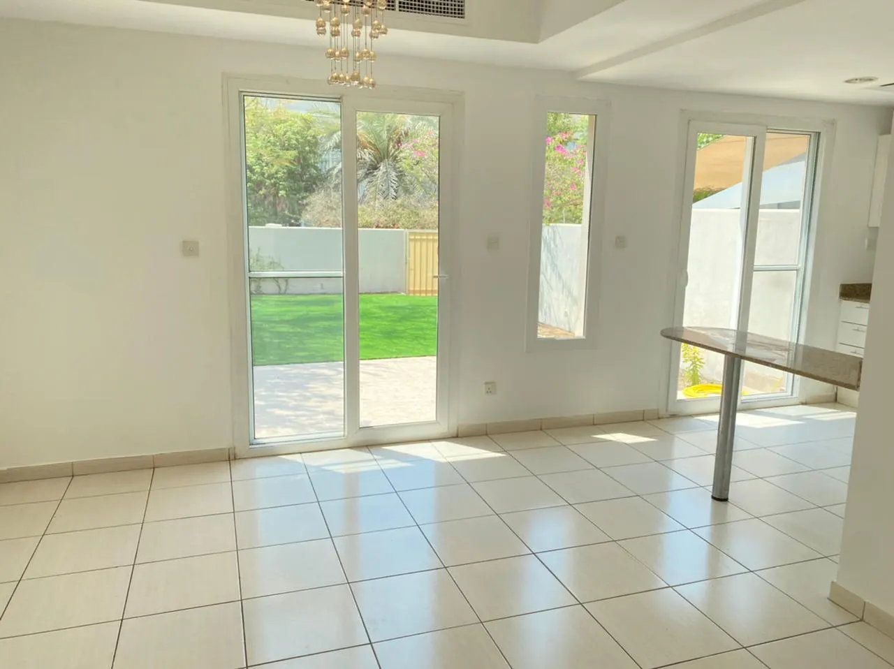 Bright, empty room with white walls, tile floor, and sliding glass doors to a green lawn. A chandelier hangs from the ceiling.