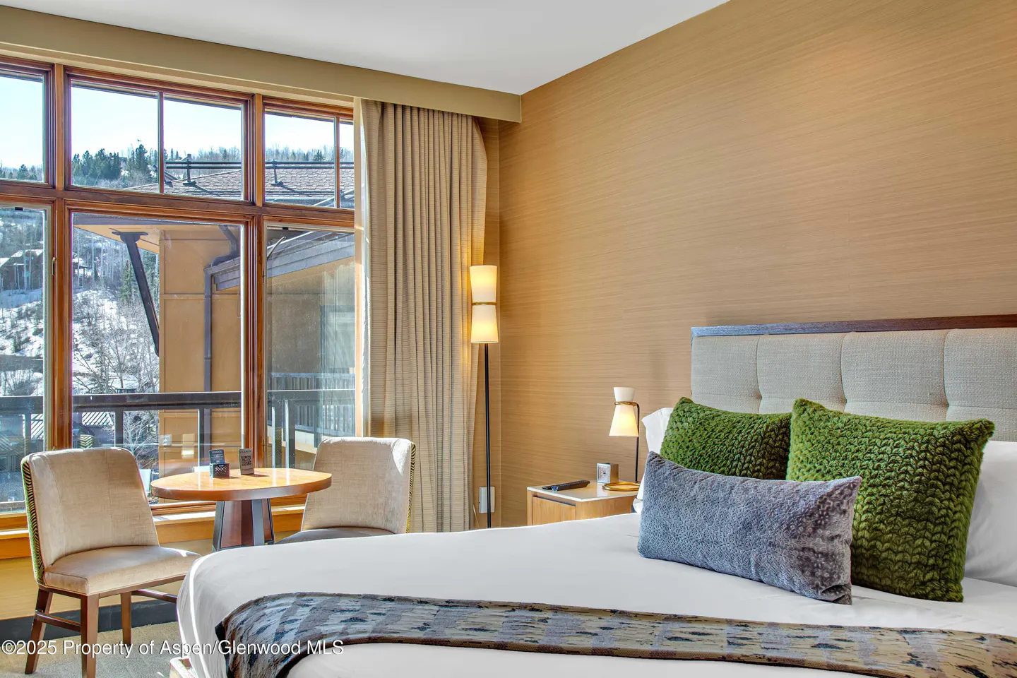 Bedroom with a king bed, green pillows, and a view of a snowy mountain through a large window. There is a table and two chairs by the window.