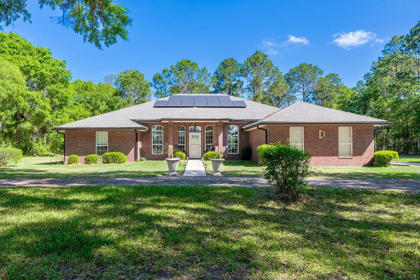 Brick ranch-style home with solar panels on the roof, a white front door, and a green lawn on a sunny day.