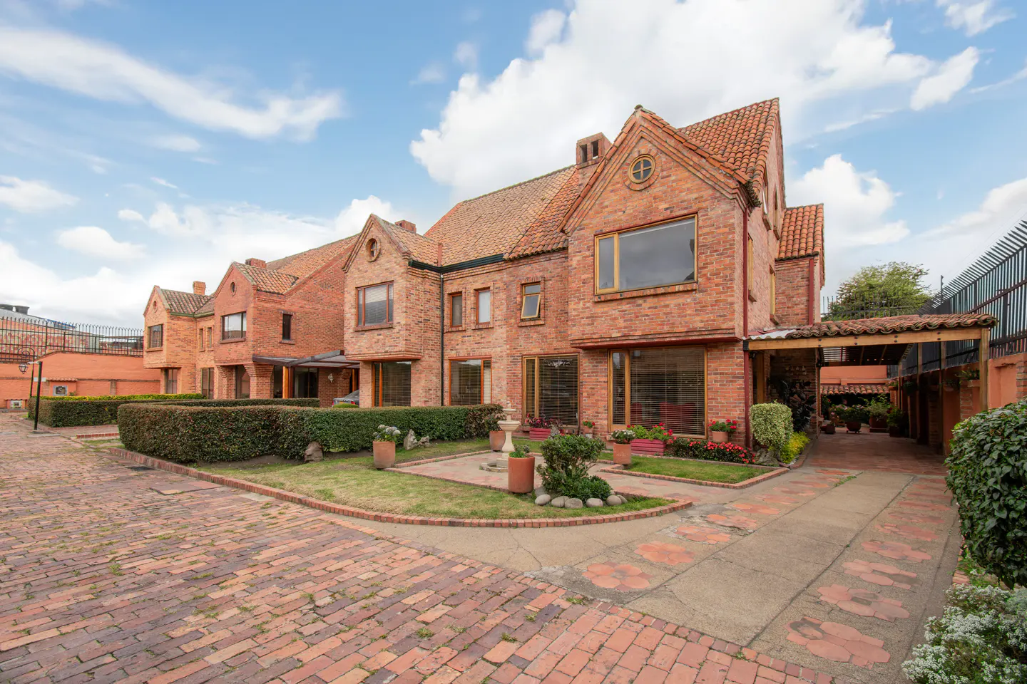 Exterior view of red brick townhouses with red tile roofs, green hedges, and a brick driveway under a cloudy sky.