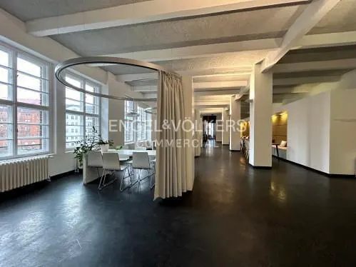 Bright, open commercial space with black floors, white walls, and large windows. A table with white chairs sits near the windows, partially enclosed by a curtain. "Engel & Völkers Commercial" logo visible.