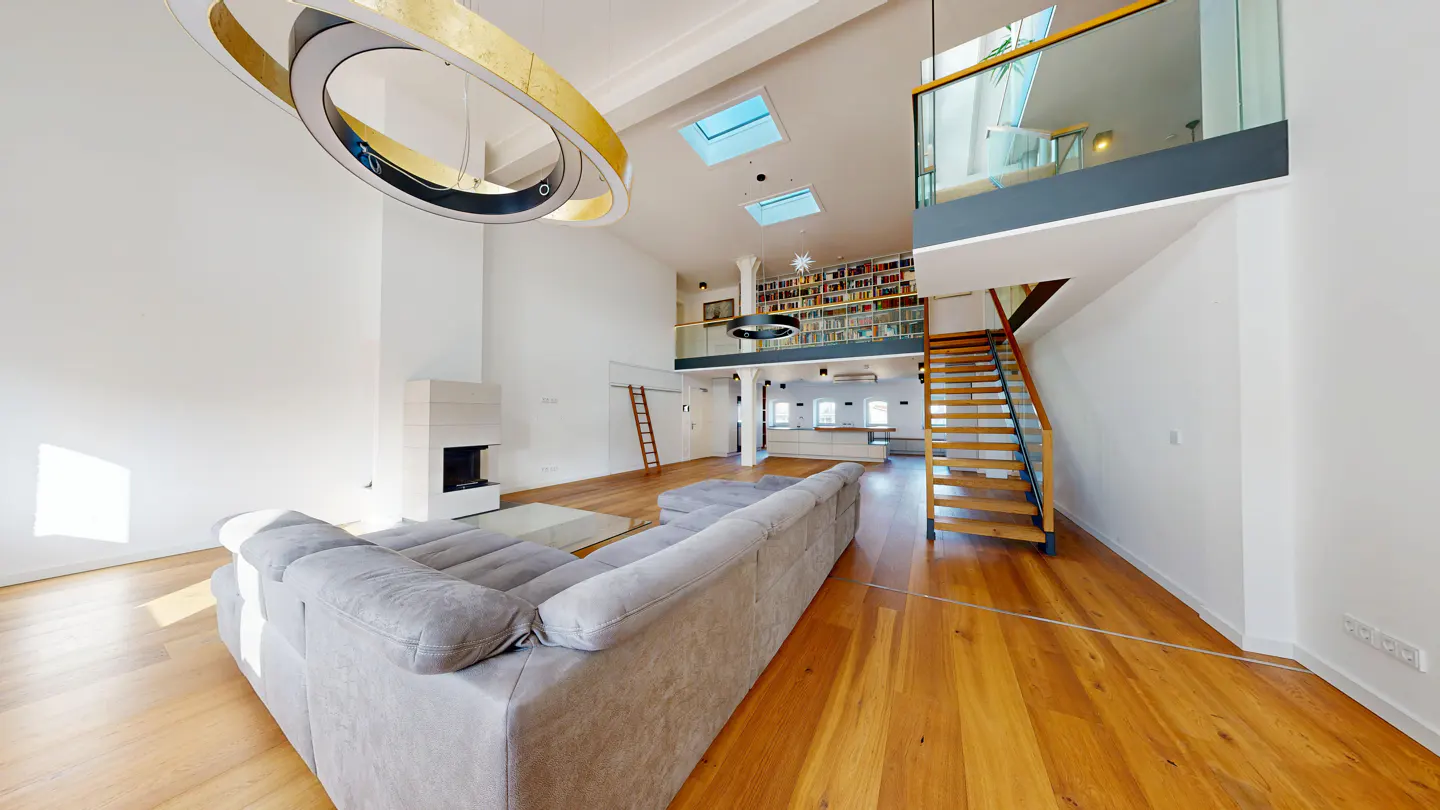 Bright, modern living room with hardwood floors, a gray sofa, and a white fireplace. A staircase leads to a loft with a bookshelf.