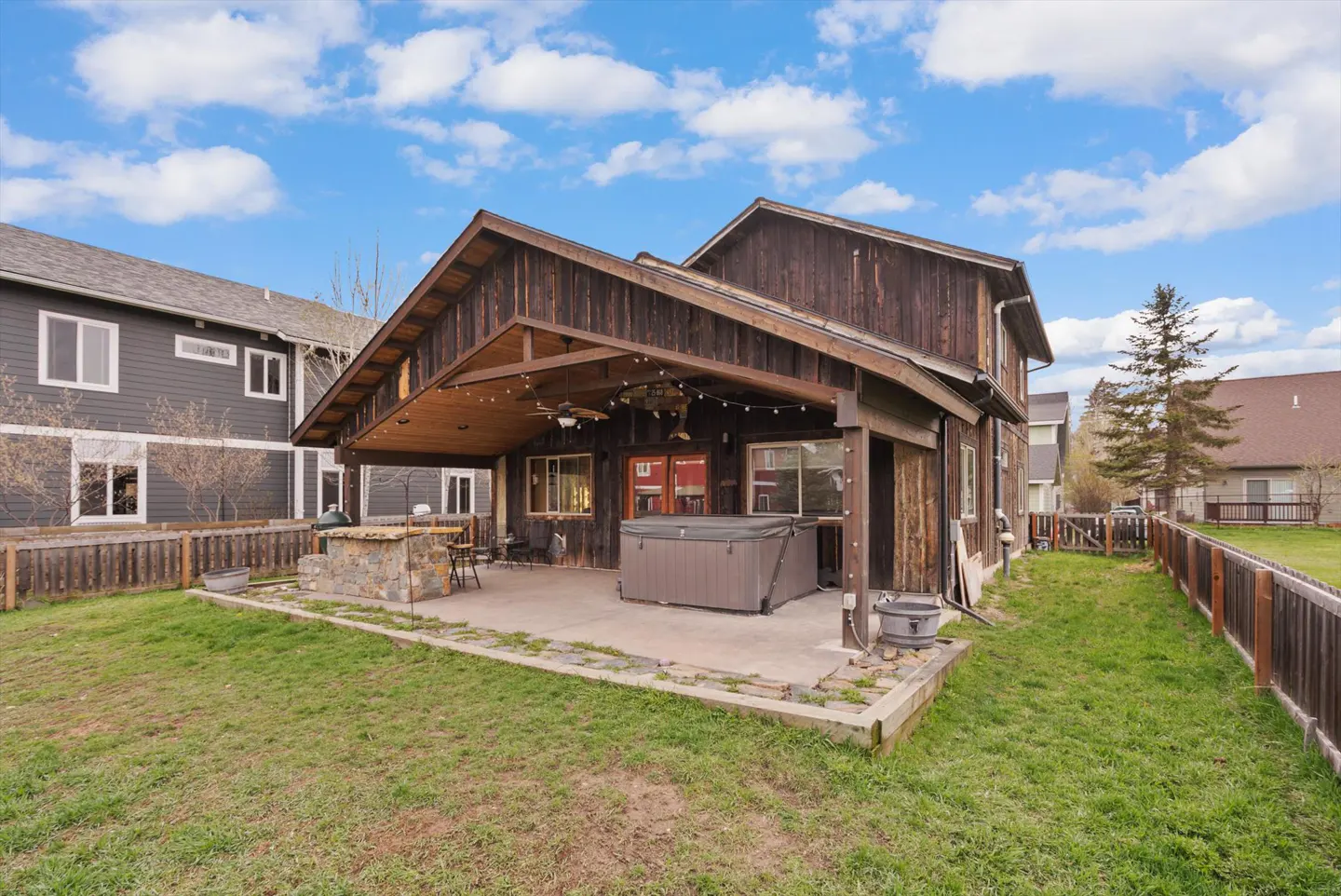 Backyard view of a brown wood house with a covered patio, hot tub, stone bar, and green lawn under a partly cloudy sky.