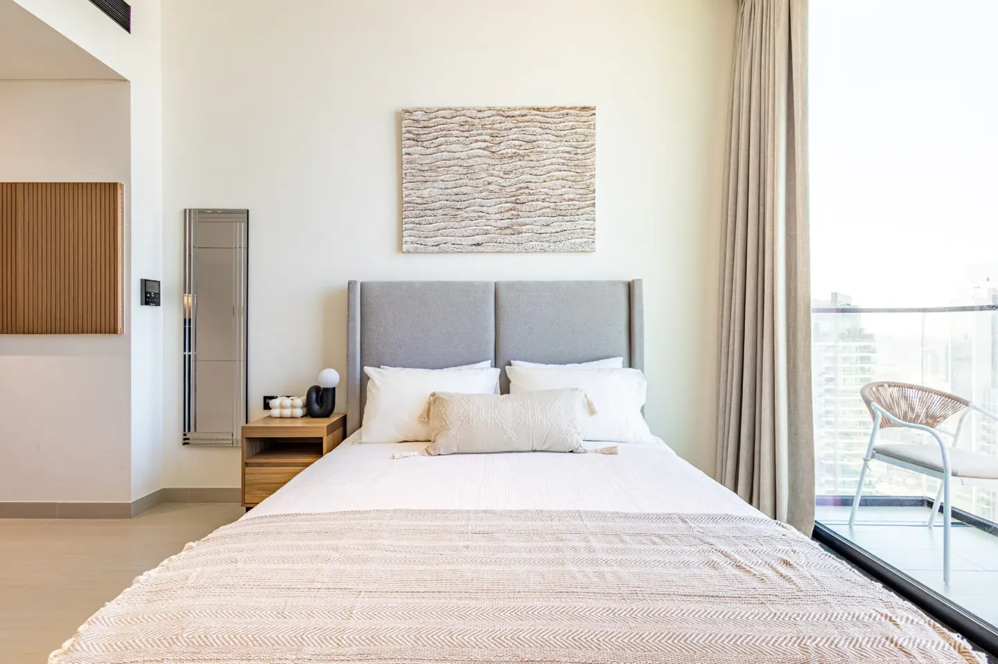 Bedroom with a gray headboard, white linens, and a beige throw blanket. A balcony with a chair is visible through the window.