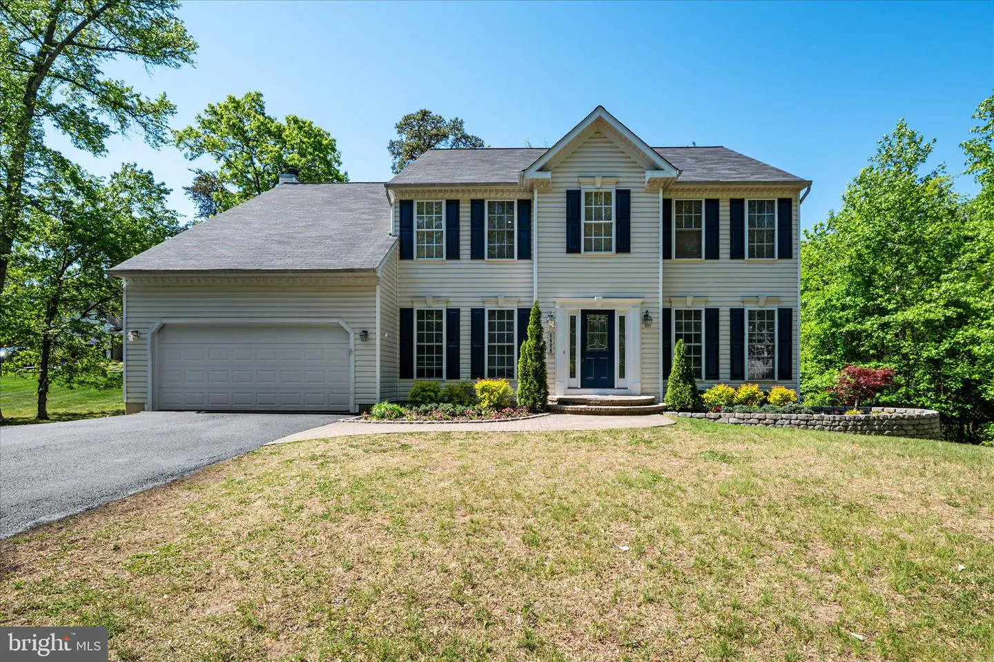 Two-story beige house with black shutters, a gray roof, and a two-car garage on a sunny day.