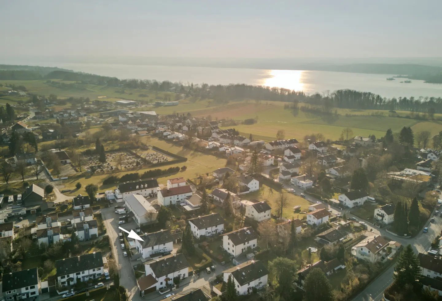 Aerial view of a village with white houses, green fields, trees, and a lake in the background. An arrow points to a house.
