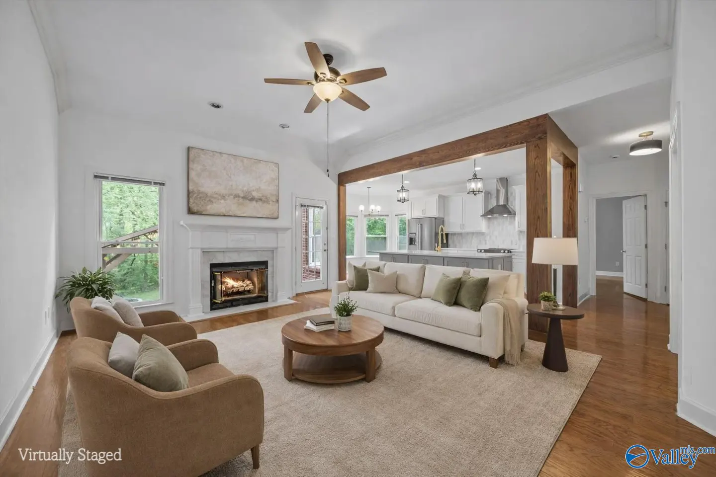 Open-concept living room with a fireplace, beige sofa, and two armchairs. A wood beam separates the living room from the kitchen.