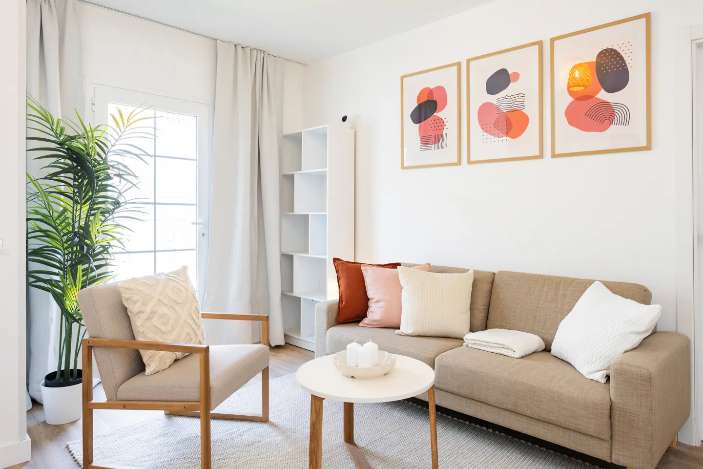 Living room with beige sofa, chair, and white walls. Three framed abstract art pieces hang above the sofa. A potted plant sits near a window.