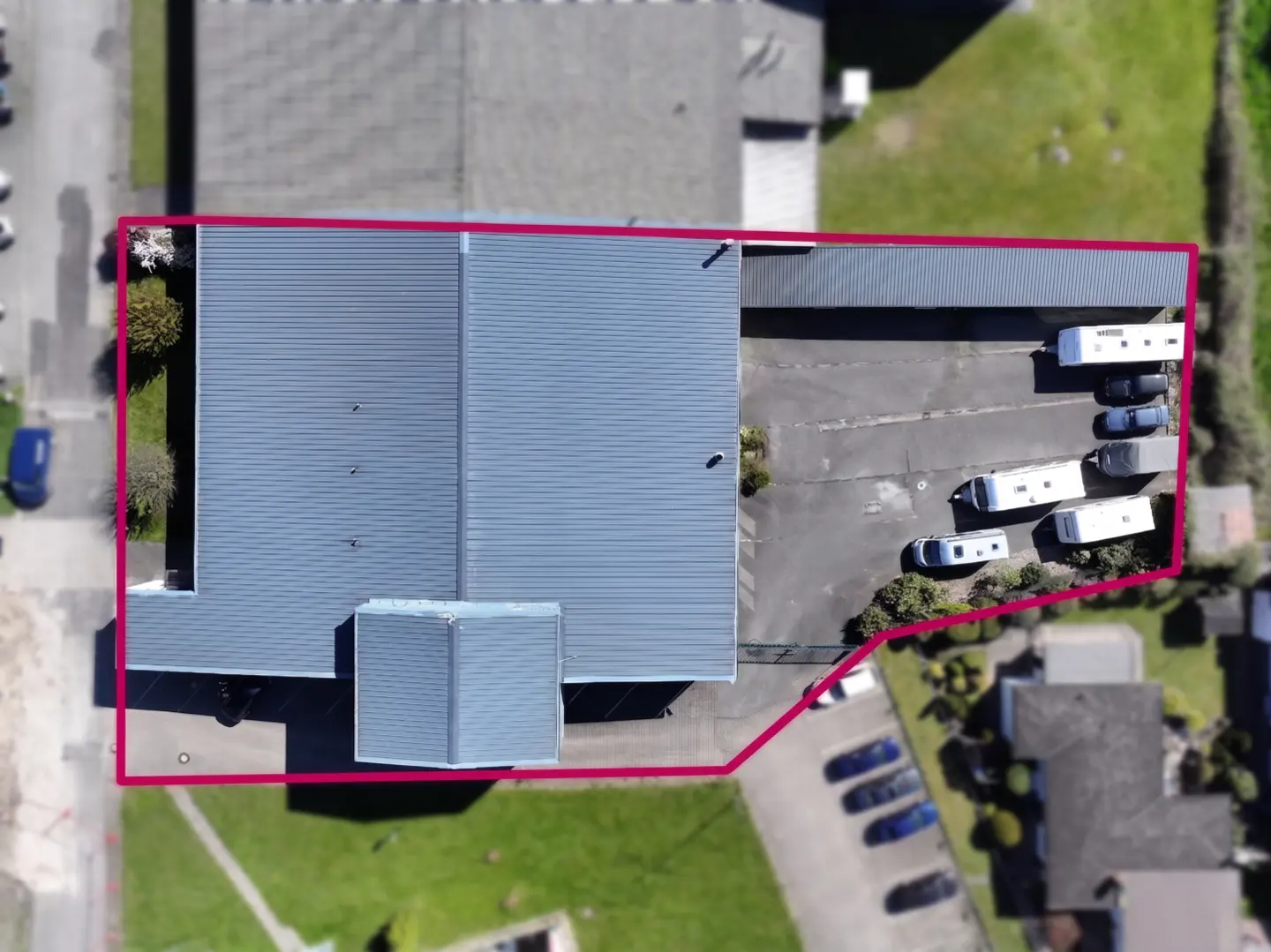 Aerial view of a commercial building with a gray roof, surrounded by a pink border, parking lot, and green spaces.