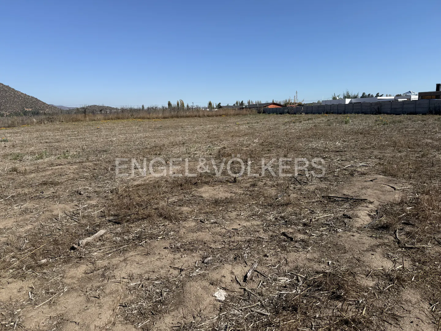 A wide shot of a vacant lot with dry grass and dirt under a clear blue sky. A mountain is visible in the distance. The Engel & Volkers logo is superimposed.