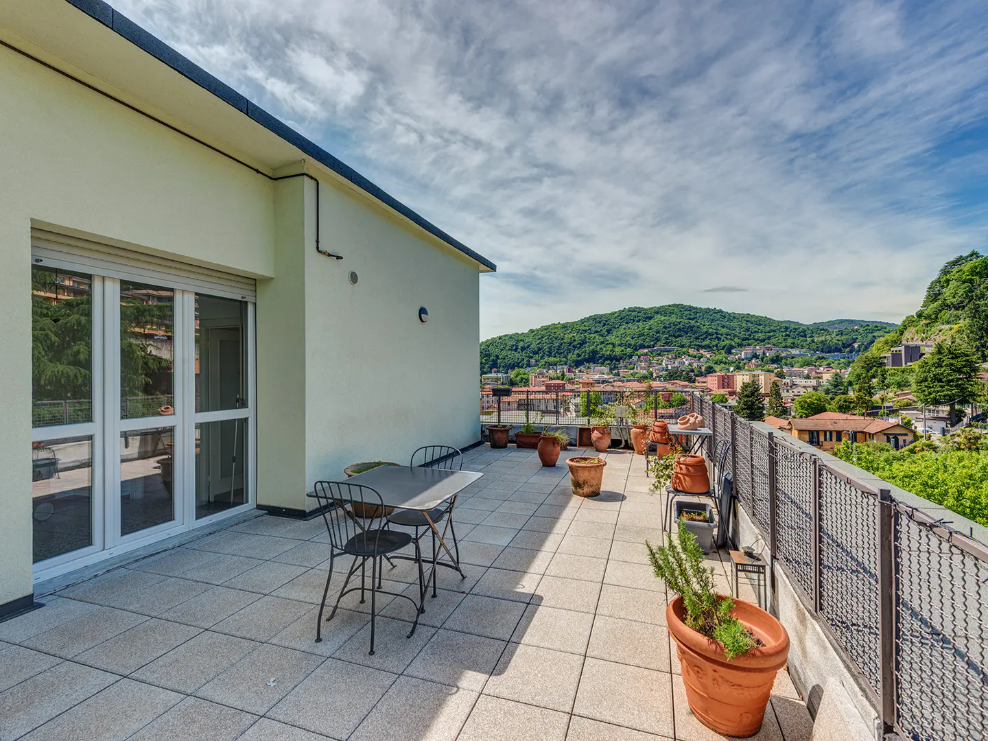 A rooftop terrace with a table, chairs, and potted plants overlooks a cityscape with green hills under a cloudy sky.