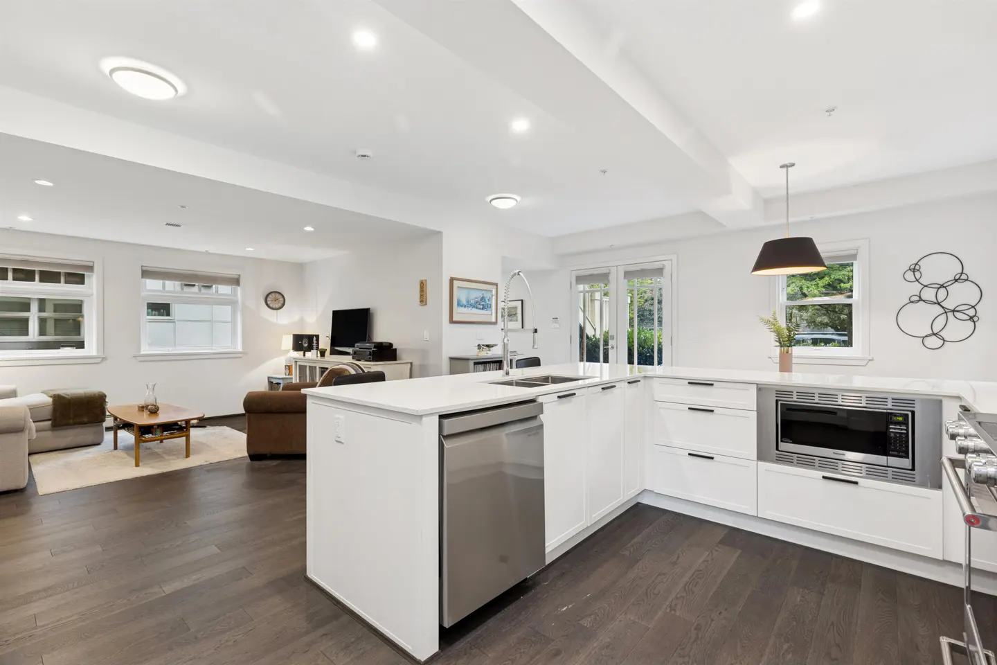 Open-concept living space with white kitchen cabinets, stainless steel appliances, and dark wood floors. Living room with sofa and TV in background.