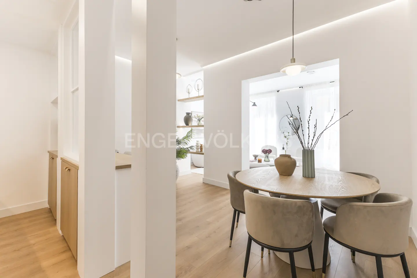 Bright, modern dining room with a round wood table and four beige chairs. Vases sit on the table. White walls and wood floors.