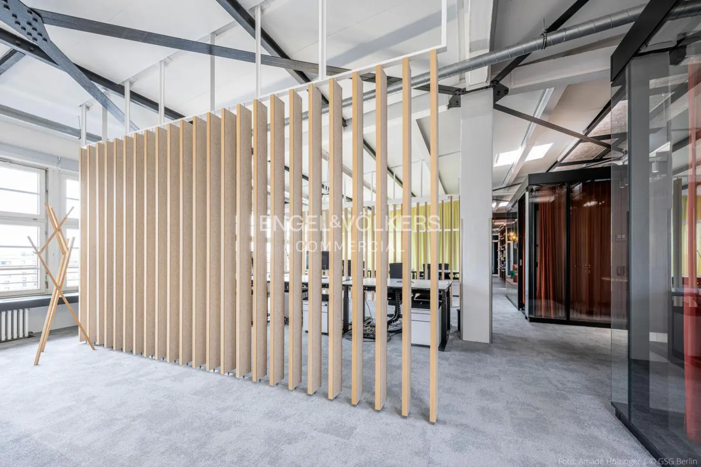 Bright office space with a light wood slat partition, grey carpet, and white ceiling with exposed beams. Desks and office furniture are visible in the background.