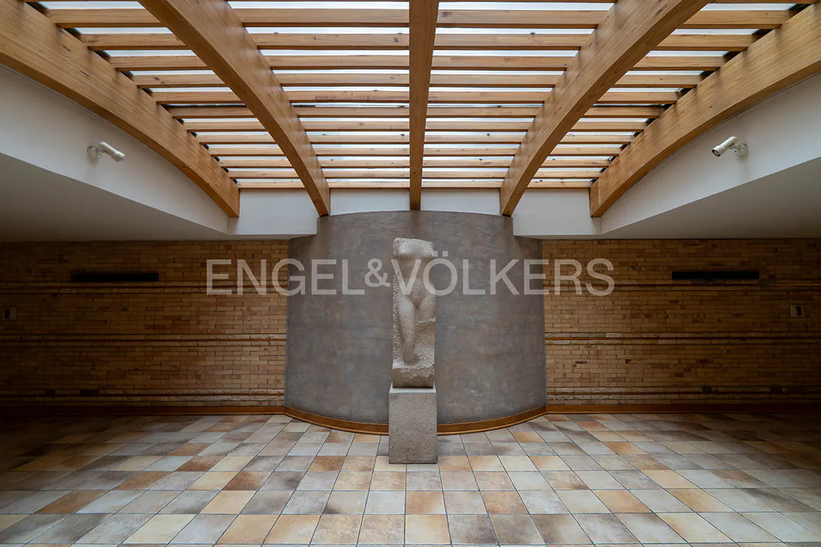 A stone sculpture stands before a curved wall in a room with a wooden slatted ceiling and tiled floor.