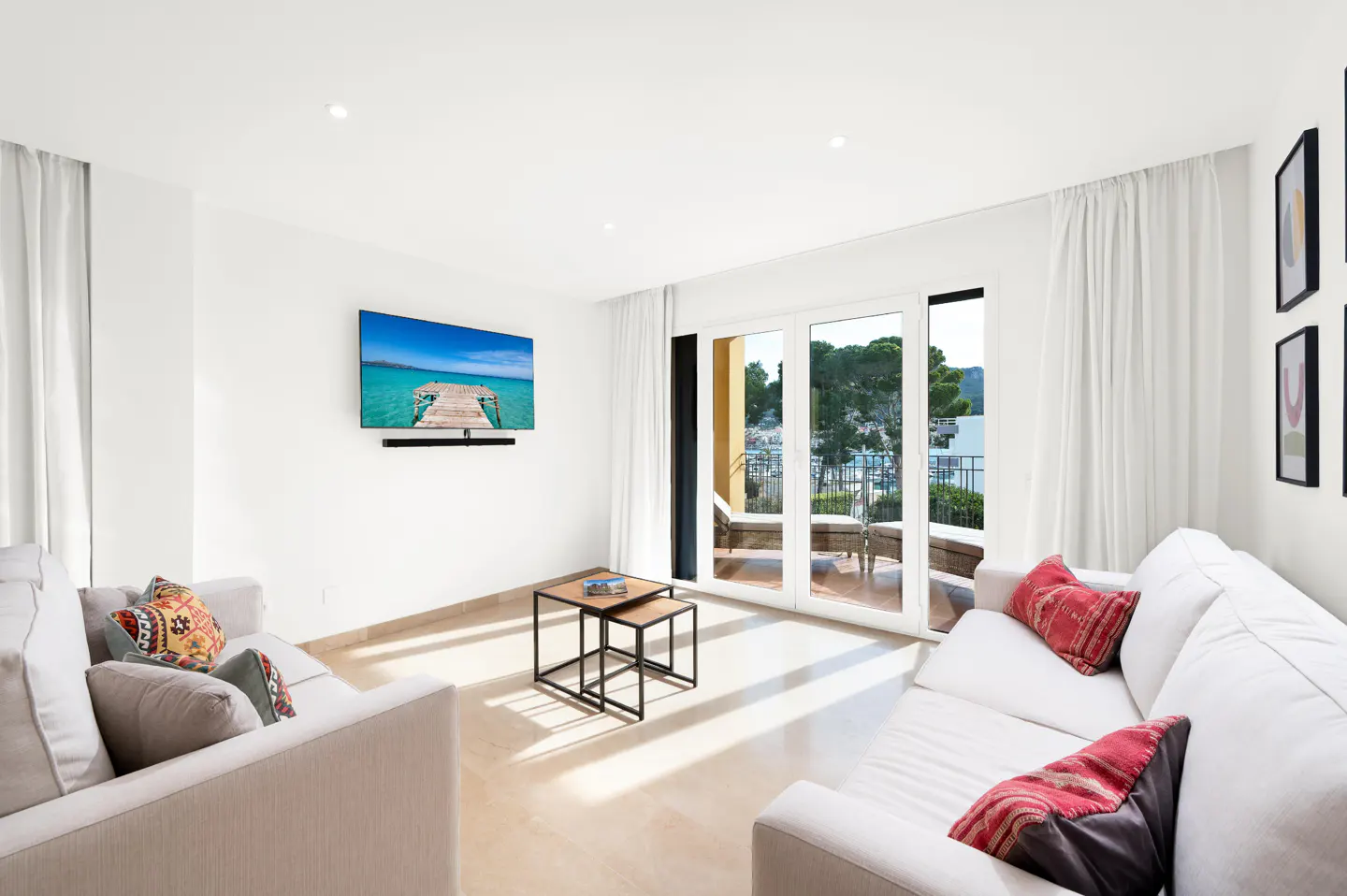 Bright living room with white sofas, red pillows, and a TV showing a beach scene. Balcony doors open to a view of trees and water.