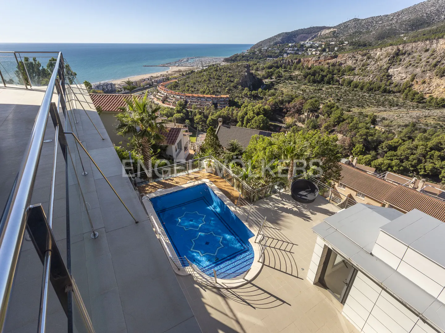 Aerial view of a blue tiled pool with star design, overlooking the ocean, beach, and mountains. Balcony with metal railing in the foreground.