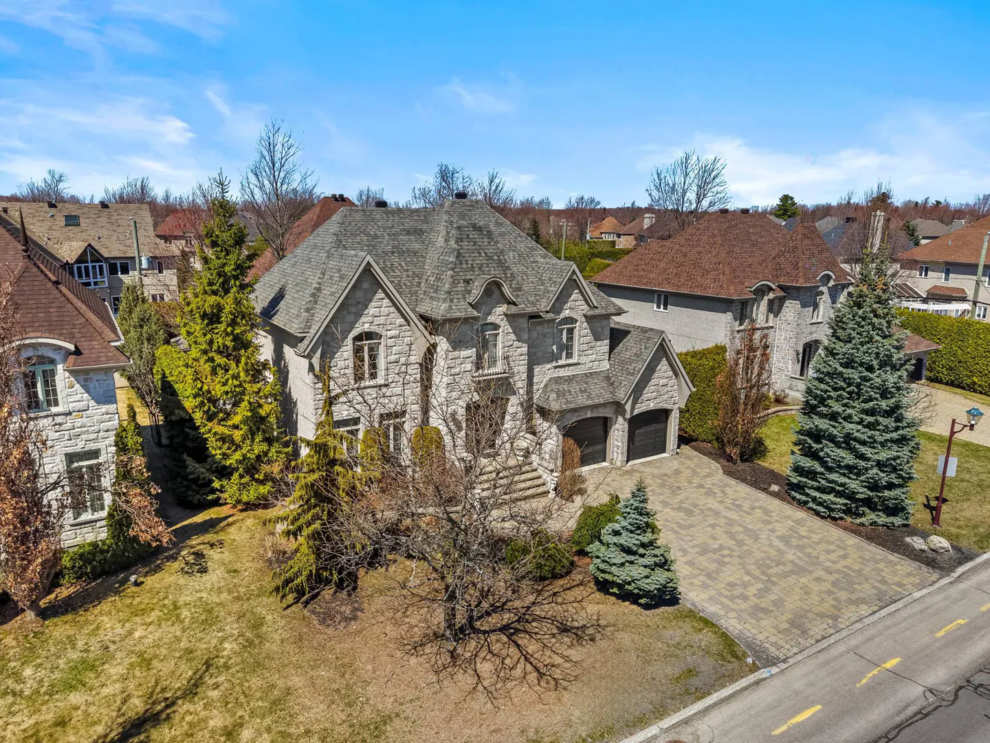 Aerial view of a large, two-story stone house with a gray roof and a paved driveway on a sunny day.