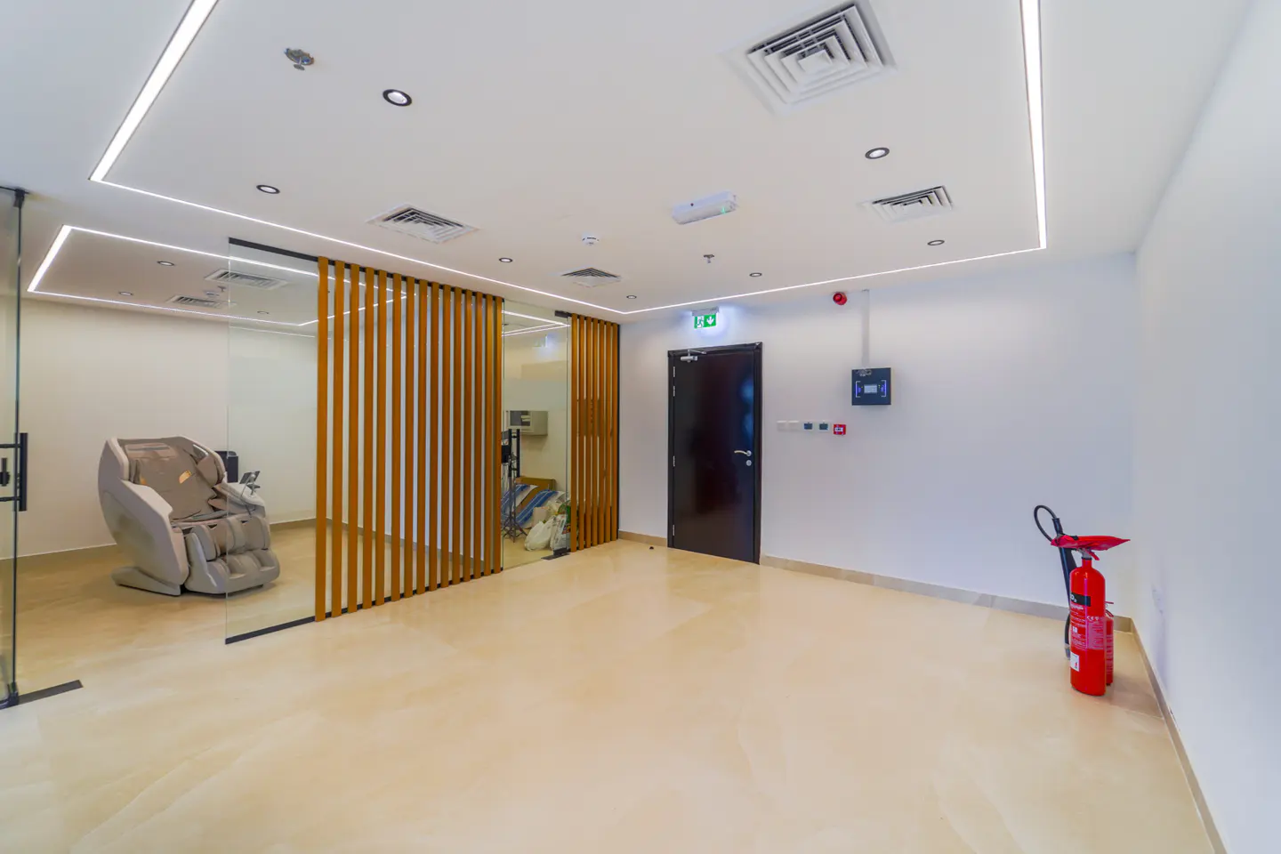 Bright room with a massage chair visible through a glass wall and wooden slats. A fire extinguisher stands near a dark door on the right.