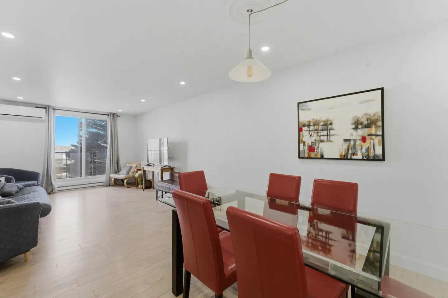 Modern dining area with a glass table and four red chairs, adjacent to a living room with a grey sofa and balcony view.