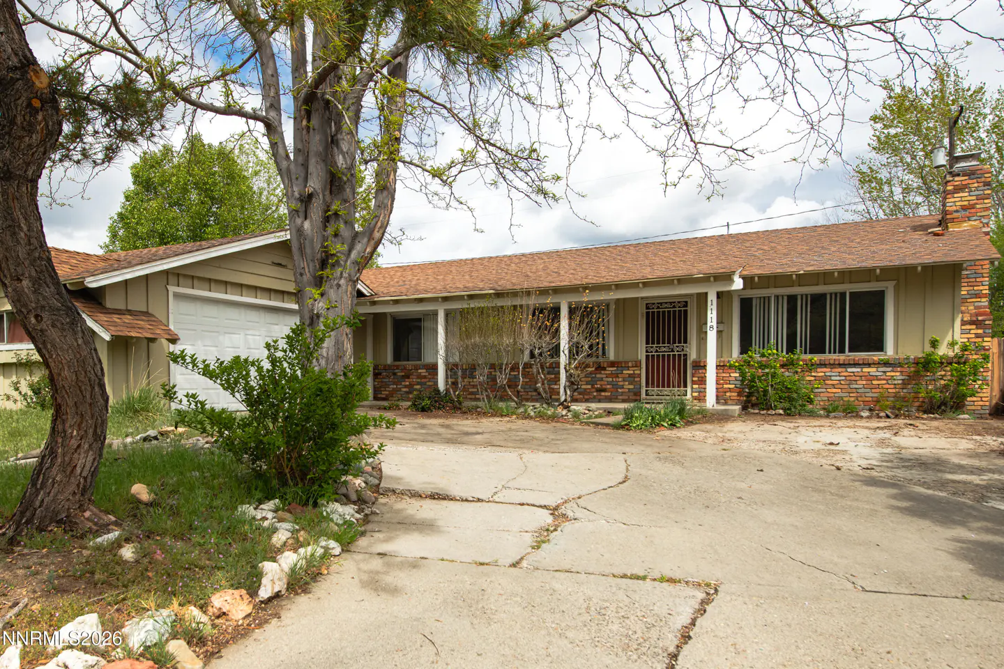 A single-story, light-yellow house with a brown roof and brick accents, featuring a cracked concrete driveway and trees.