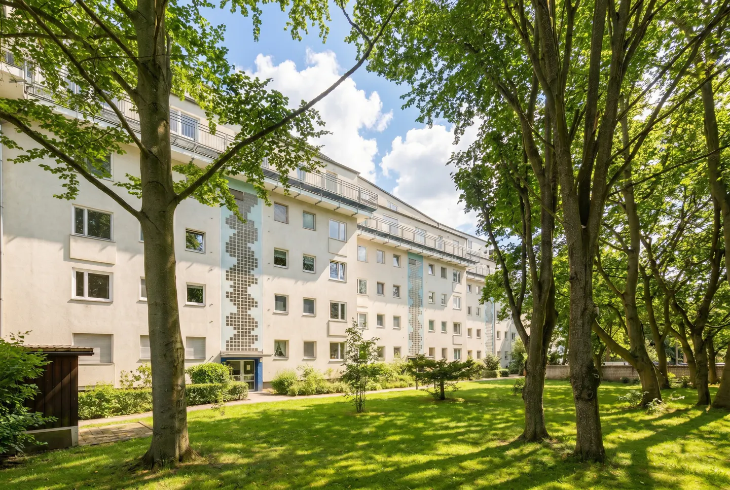 Exterior view of a multi-story apartment building with a green lawn and trees in the foreground. The building is light beige with blue accents.