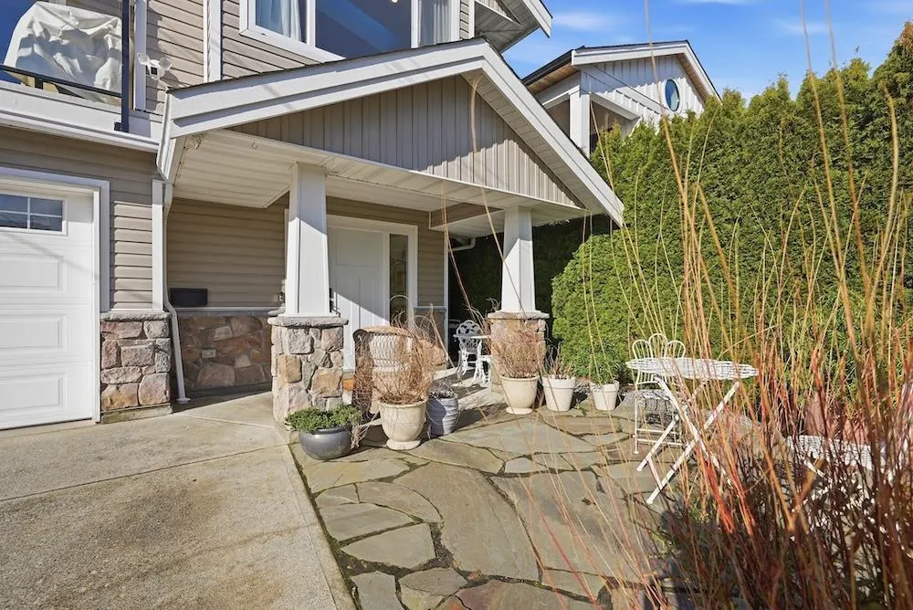 Tan house with stone accents, white trim, and a stone patio with white metal furniture and potted plants.