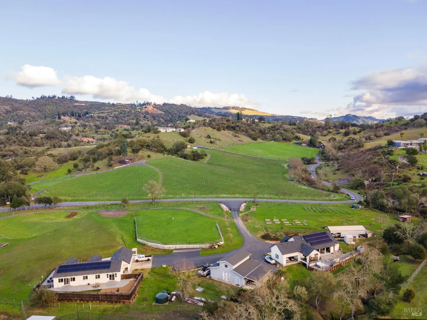 Aerial view of a vineyard estate with green fields, houses, and rolling hills under a blue sky.