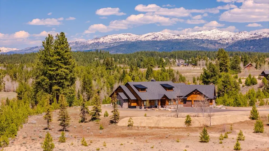 A large, wood and stone house with a gray roof sits in a clearing surrounded by trees, with snow-capped mountains in the background.