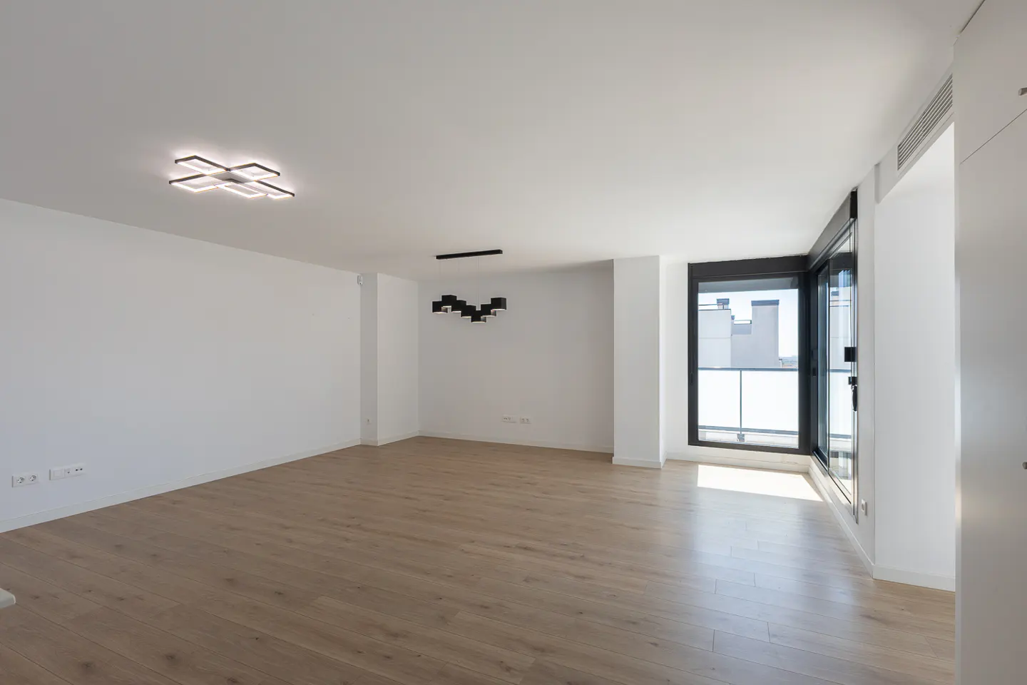 Bright, empty room with light wood floors, white walls, and a large window. Modern light fixtures on the ceiling.