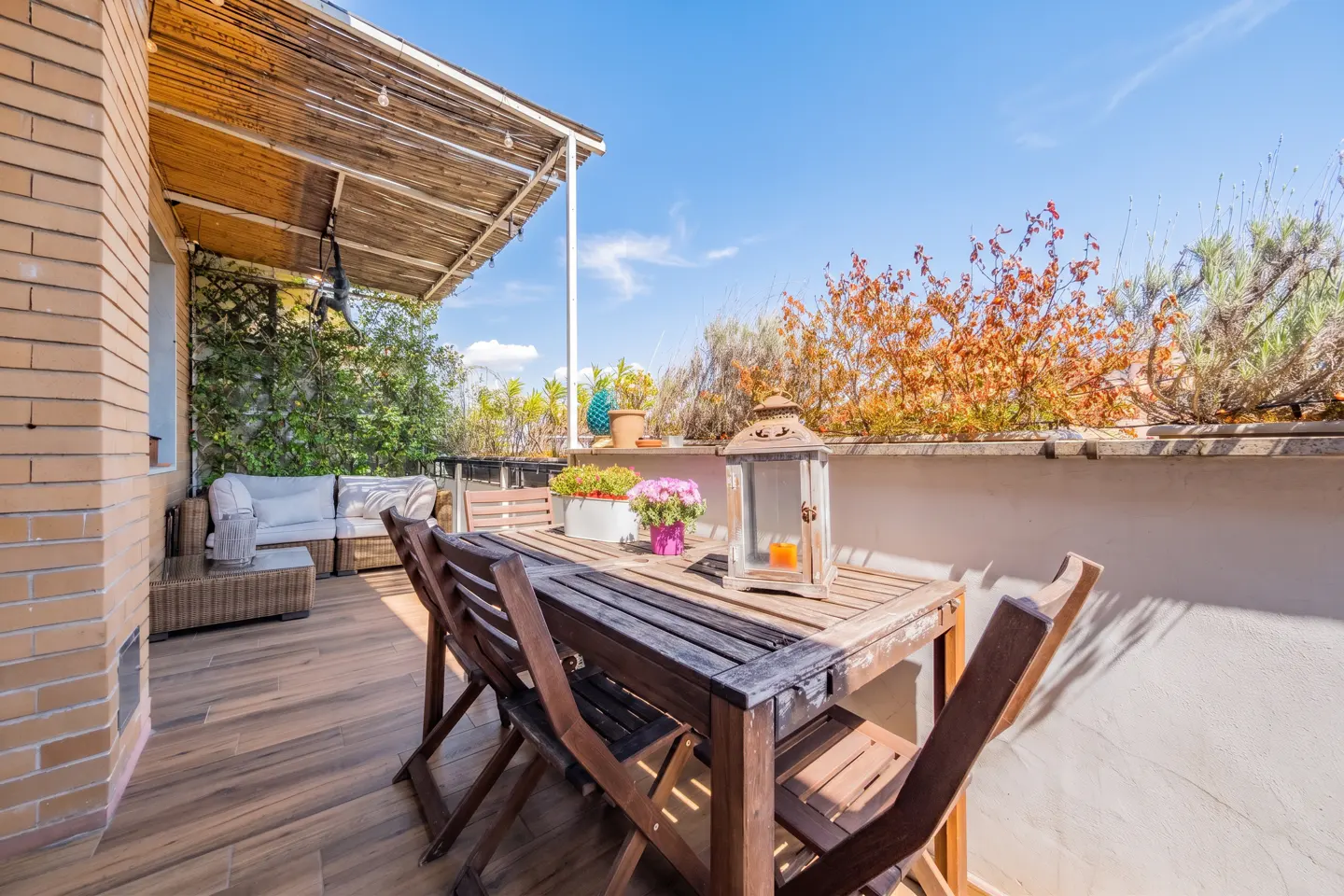 Sunny rooftop terrace with a wooden dining set, a lantern, potted flowers, and a cozy wicker sofa under a bamboo canopy.