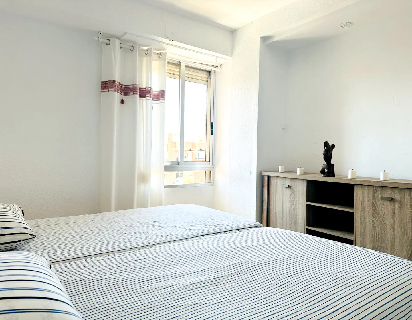 Bright bedroom with two twin beds, striped bedding, white walls, and a light wood cabinet with a dark sculpture. Window with white curtains.